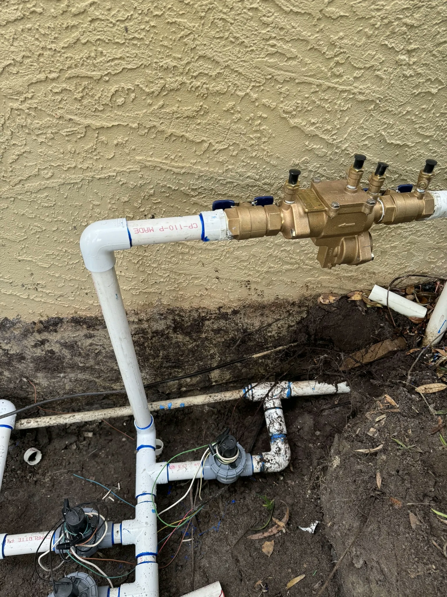 A residential sprinkler system PVC pipe installation with a brass manifold and electrical wires.