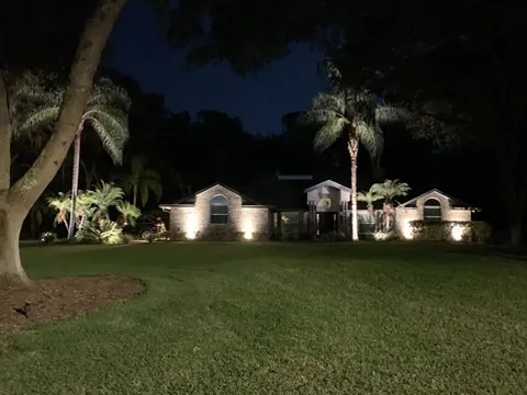 A well-lit landscape with a house, palm trees, and green lawn.